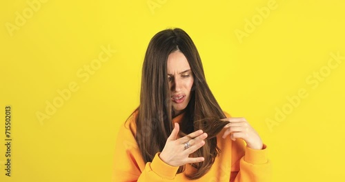 Depressed young woman looking at split brunette hair ends. Worried sad girl feels upset about brittle damaged dry hair loss concept. Female hormone problems concept isolated yellow background. photo