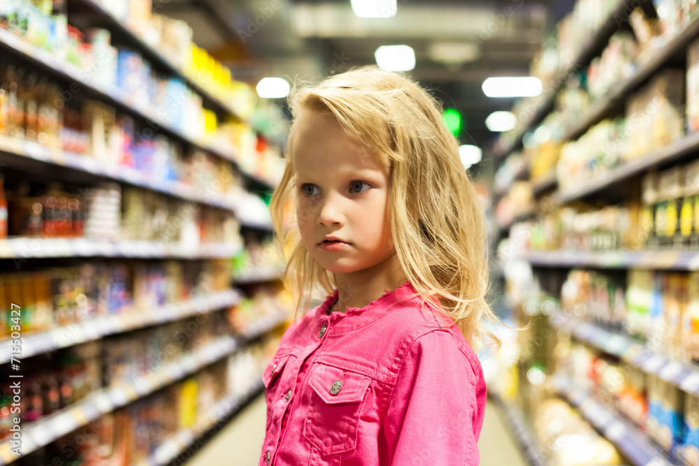 Child girl 5 year old at shelves with groceries in store background, bored looking away. Portrait of cute kid girl shopping choice buying in supermarket. Retail shopping concept. Copy ad text space