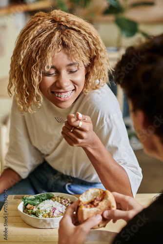cheerful african american woman with braces eating salad near curly boyfriend with tofu burger