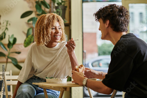 happy african american woman with braces eating salad bowl near blurred boyfriend in vegan cafe photo