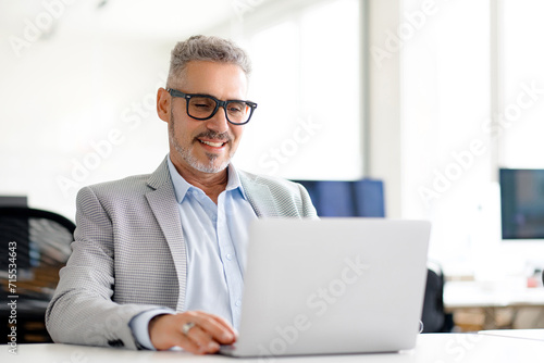 Mature handsome grey-haired businessman in a smart blazer working diligently on his laptop, reflecting the focus and engagement that characterize today's business leaders