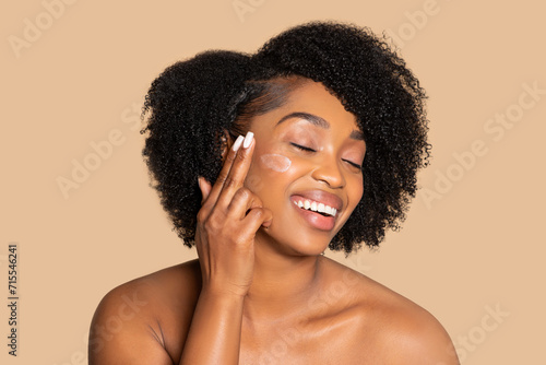 Joyous woman applying moisturizer on cheek, beauty routine photo