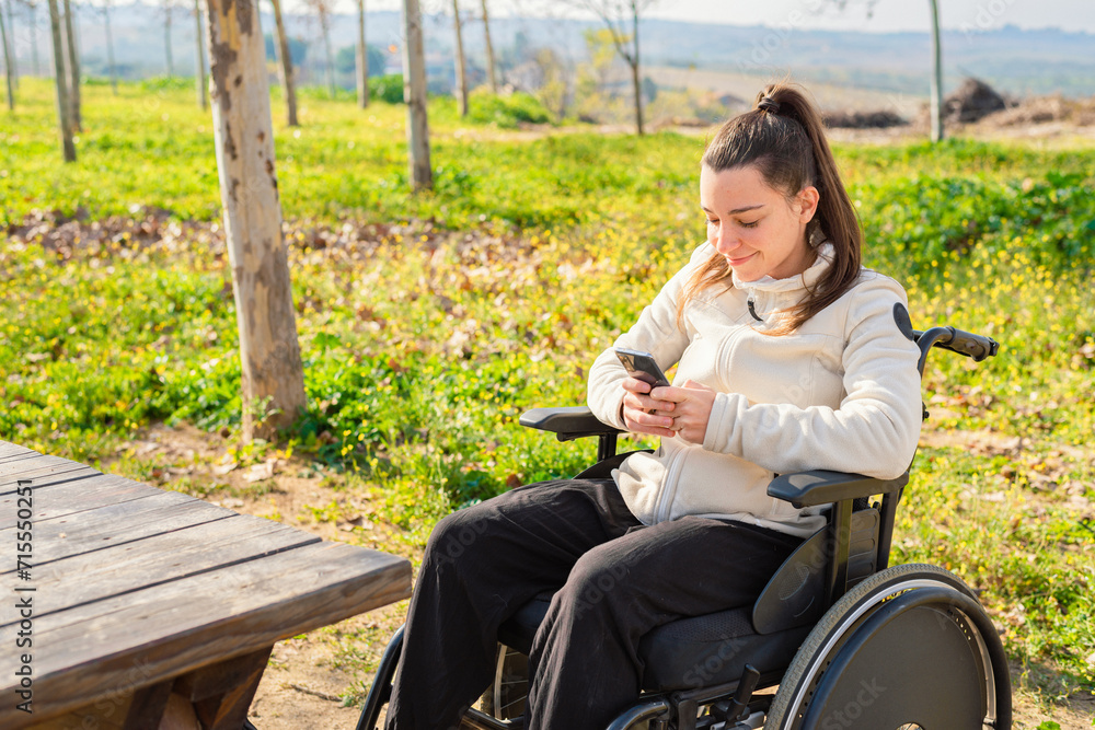 A young woman in a wheelchair is smiling while browsing on her smartphone in a park A young woman in a wheelchair is smiling while browsing on her smartphone in a park