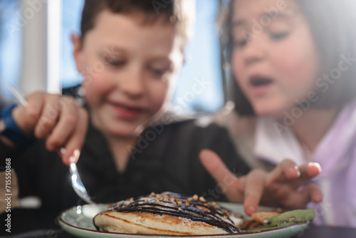 Two children enjoying a meal together, with a boy holding a fork over a pancake topped with syrup and a girl pointing towards it photo