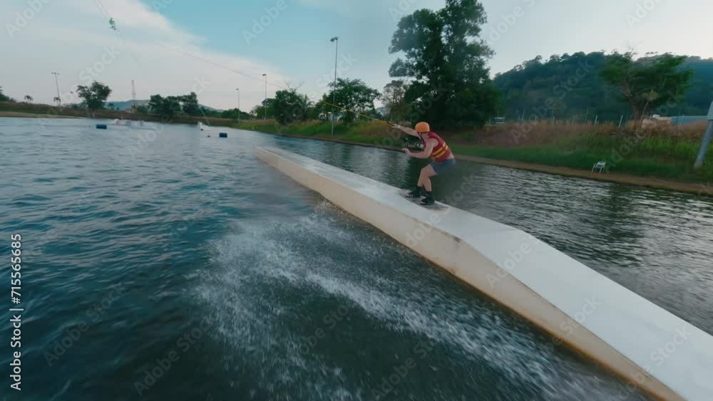 Beginner wakeboarder man in orange helmet trying easy tricks on cable wakeboard lake park water sport. Extreme activity athlete guy training practicing wakeboarding water sport fpv drone follow shot Beginner wakeboarder man in orange helmet trying easy tricks on cable wakeboard lake park water sport. Extreme activity athlete guy training practicing wakeboarding water sport fpv drone follow shot