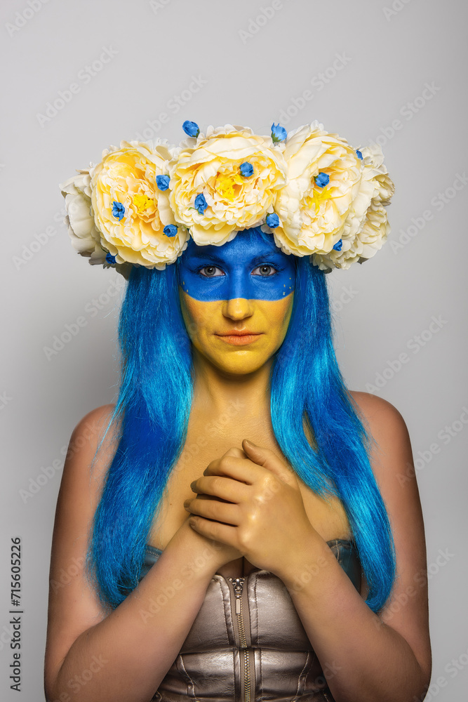 Young woman with yellow blue face art, wreath of peonies on head, praying for salvation on an isolated gray background. Creative concept for Flag Day in Ukraine. Copy space. Girl looking at camera Young woman with yellow blue face art, wreath of peonies on head, praying for salvation on an isolated gray background. Creative concept for Flag Day in Ukraine. Copy space. Girl looking at camera