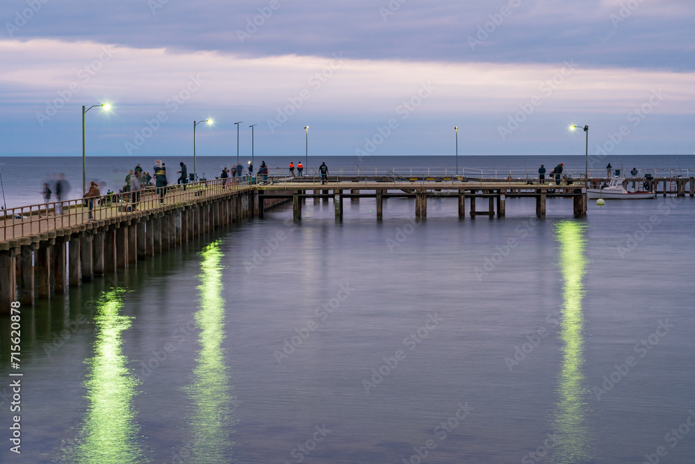 Fishermen and tourists on wooden pier over a calm coastal bay at twilight Fishermen and tourists on wooden pier over a calm coastal bay at twilight