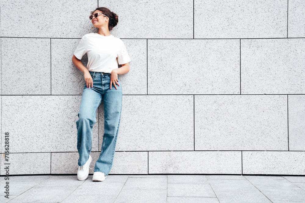 Young beautiful smiling hipster woman in trendy summer white t-shirt and jeans clothes. Carefree woman, posing in the street at sunny day. Positive model outdoors near wall. In sunglasses Young beautiful smiling hipster woman in trendy summer white t-shirt and jeans clothes. Carefree woman, posing in the street at sunny day. Positive model outdoors near wall. In sunglasses