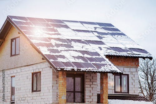 Construction Eco House with Solar Panels on Roof in Winter. New Low Energy Home with Snow Covered Photovoltaic Solar Panels on Roof under Construction. 