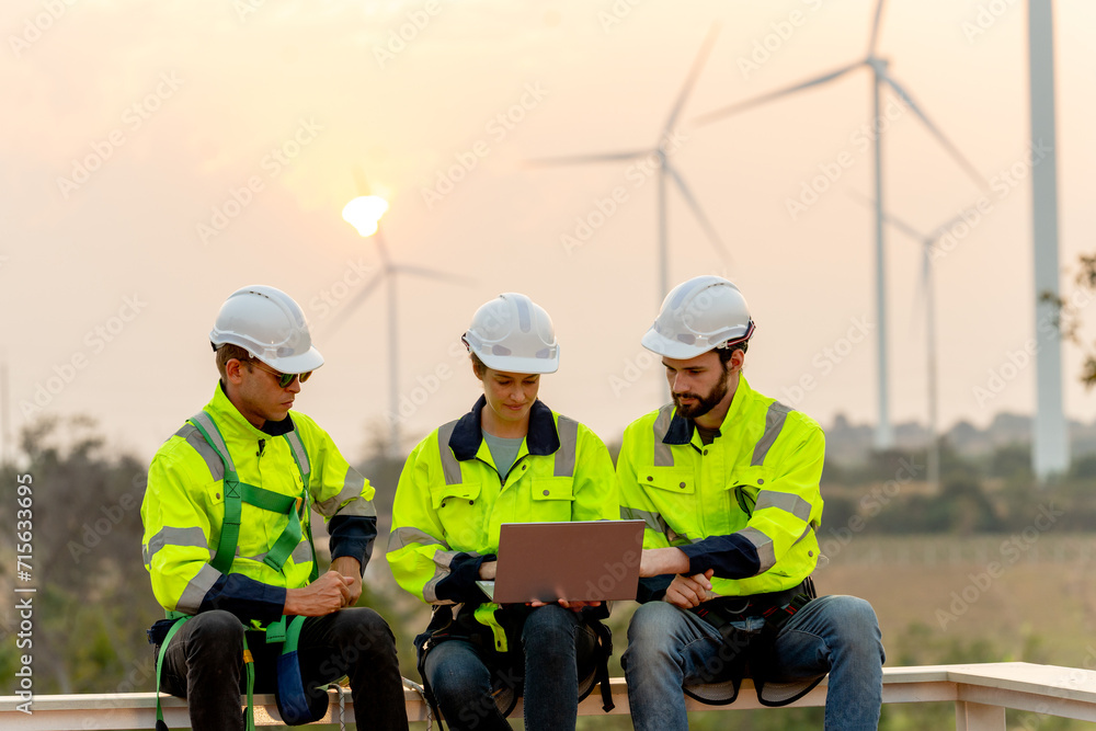 Close up group of professional technician workers with safety suit sit and discuss work using laptop and row of windmill or wind turbine on the background in evening. Close up group of professional technician workers with safety suit sit and discuss work using laptop and row of windmill or wind turbine on the background in evening.