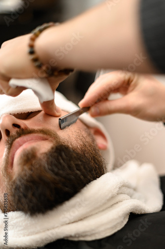 A barber shaves the cheek of a bearded customer with dangerous razor. Shaving the contour of the beard for the correct shape.