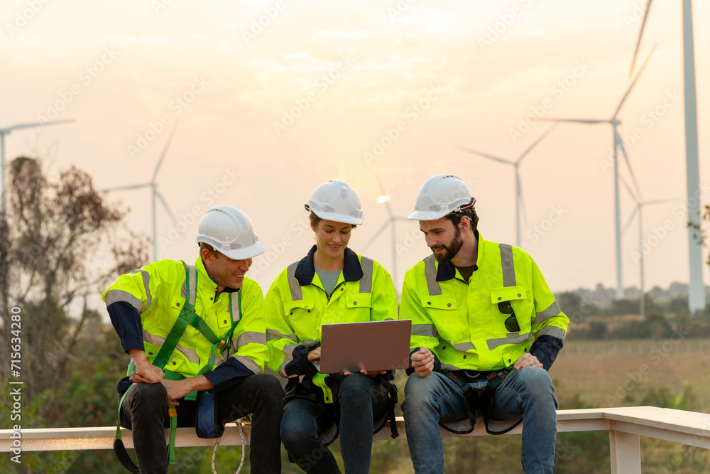 Group of professional technician workers with safety suit sit and discuss work using laptop and row of windmill or wind turbine on the background in evening. Group of professional technician workers with safety suit sit and discuss work using laptop and row of windmill or wind turbine on the background in evening.