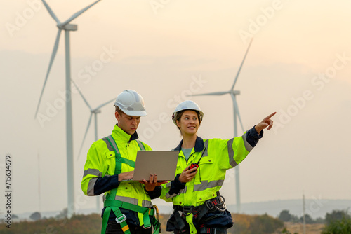 Portrait technician workers use laptop to work and stay with his co-worker point to right side in evening with windmill or wind turbine on the background.