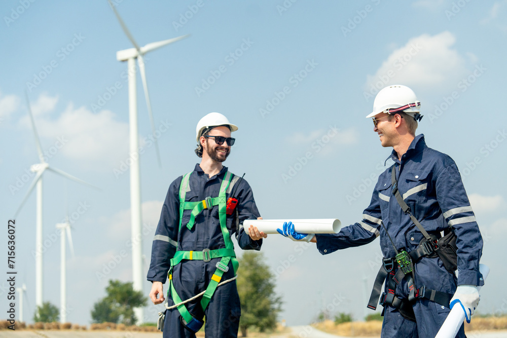 Professional Caucasian workers or technician with safety suit hold and send the construction paper plan to co-worker also stand in front of wind turbine or windmill in the row as power plant business. Professional Caucasian workers or technician with safety suit hold and send the construction paper plan to co-worker also stand in front of wind turbine or windmill in the row as power plant business.