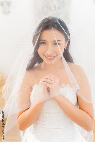A beautiful, smiling Southeast Asian bride in wedding veil while praying with rings on her finger. Wedding preparation concept.