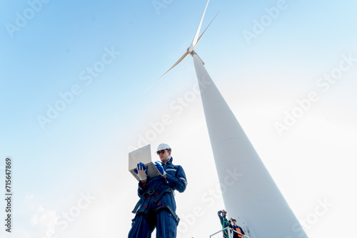 Professional technician man stand with hold laptop and stand near base of windmill or wind turbine with his co-worker in the back. photo
