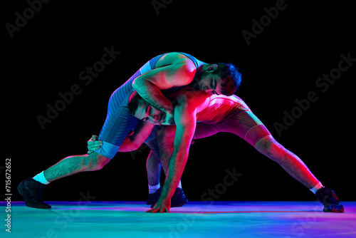 Two strong wrestlers in blue and red wrestling uniform fighting and making hip throw in motion against black background in mixed neon lights. Concept of combat sports, strength, power, movement