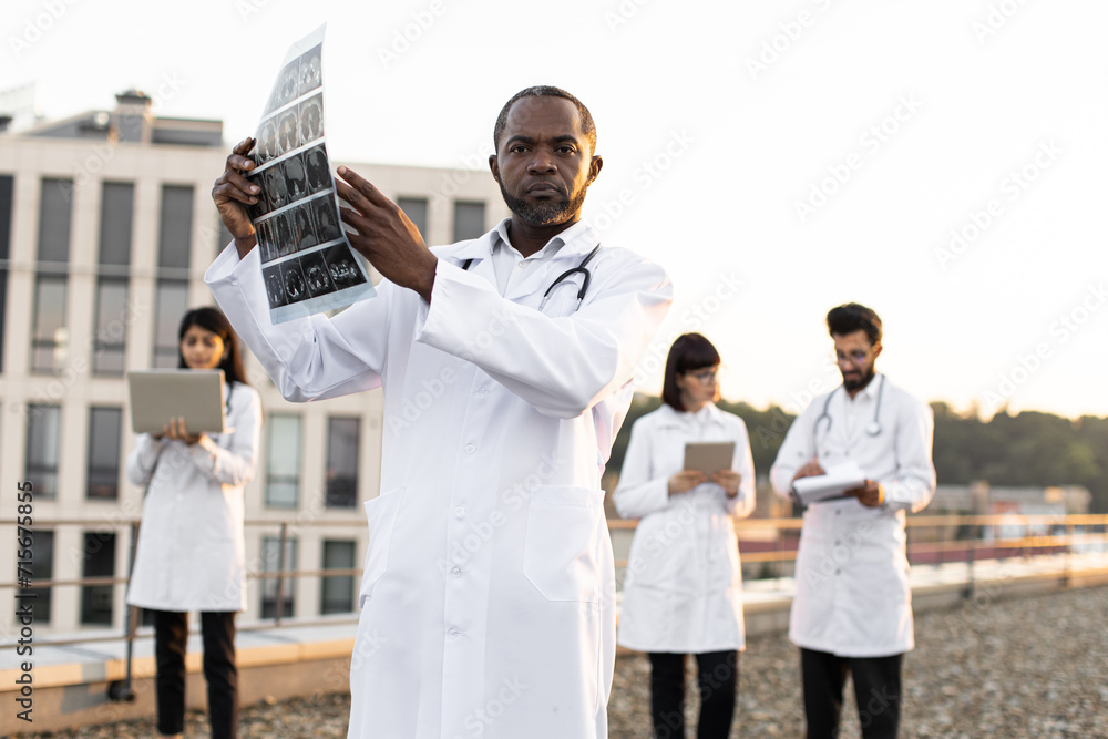 Portrait of African doctor holding x-ray scan of patient during break outdoors hospital looking at camera. Multiethnic employees of clinic working on devices with results of examination of patients. Portrait of African doctor holding x-ray scan of patient during break outdoors hospital looking at camera. Multiethnic employees of clinic working on devices with results of examination of patients.
