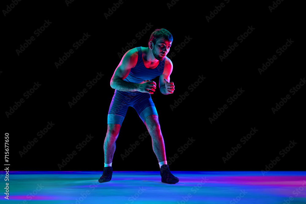 Sportsman, freestyle wrestler in blue fighting tights stands in attack position and looking ta camera against black background in mixed neon lights. Concept of professional sport, strength and power.