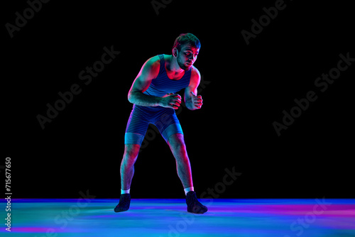 Sportsman, freestyle wrestler in blue fighting tights stands in attack position and looking ta camera against black background in mixed neon lights. Concept of professional sport, strength and power. © Lustre