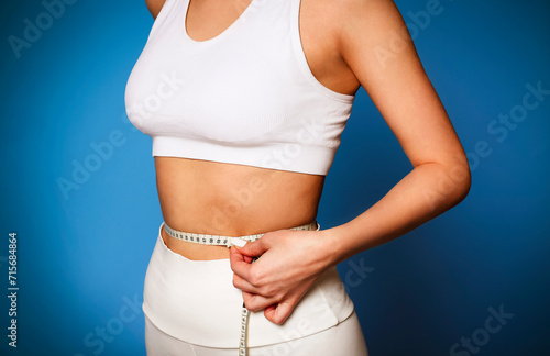 Beautiful skinny slim girl measuring her thin waist by tape measure at the blue backgriund