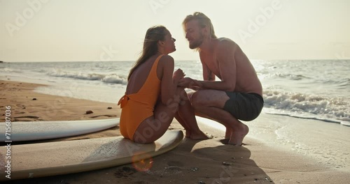 A blonde girl in an orange swimsuit sits on a surfboard, communicates with her blonde boyfriend and kisses him on the sandy beach near the sea in the morning at Sunrise photo