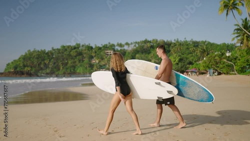 Athletic fit man, slender woman walk to ocean beach, carry surfboards. Young surfers couple dating. Boyfriend, girlfriend go surfing on sandy sea shore. Slim girl, guy with boards. Paradise coastline.