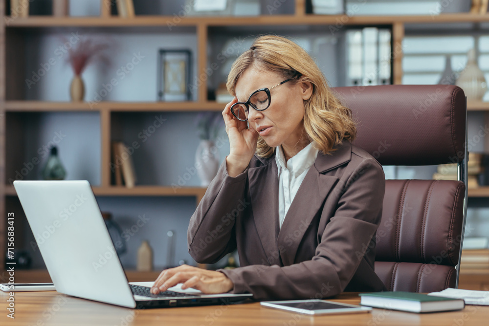 Elegant mature businesswoman concentrating as she works on a laptop from a well-appointed home office with tasteful decor. Elegant mature businesswoman concentrating as she works on a laptop from a well-appointed home office with tasteful decor.