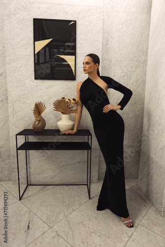 beautiful woman with dark hair in elegant black dress with accessories posing in luxurious hotel lobby