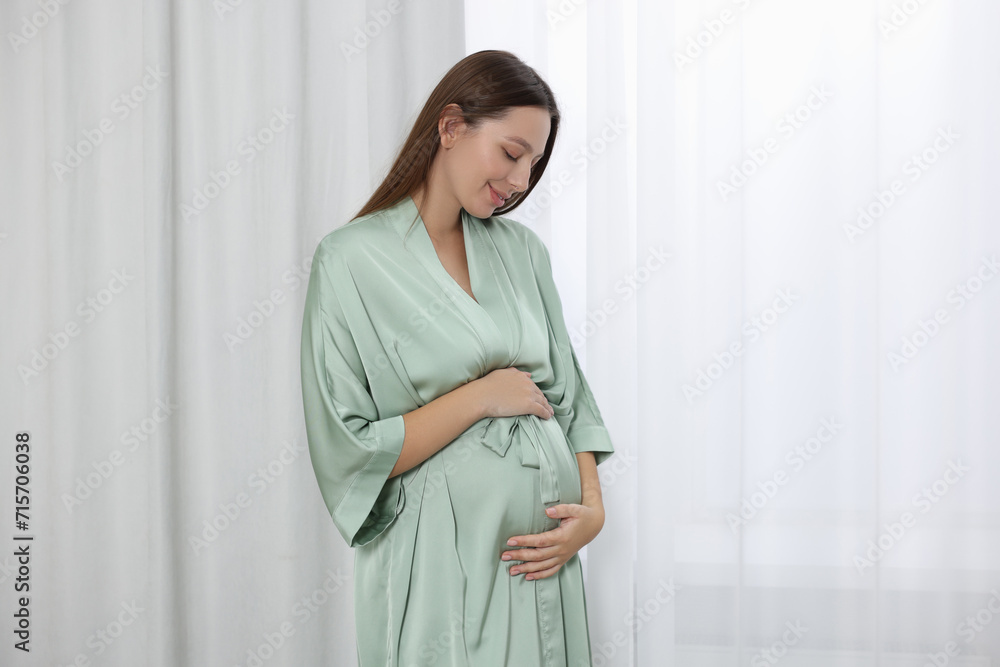Beautiful pregnant woman in green dressing gown near window indoors Beautiful pregnant woman in green dressing gown near window indoors