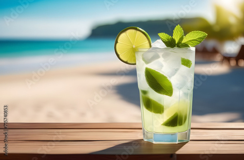 Fresh cold mojito cocktail glass with lime in a wooden table top in a beach bar, a blurred background of a sandy beach with palm trees and the azure sea. Space for text, beach holiday concept