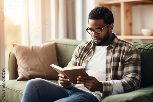 Pensive relaxed African American man reading a book at home, drinking coffee sitting on the couch. Copy space