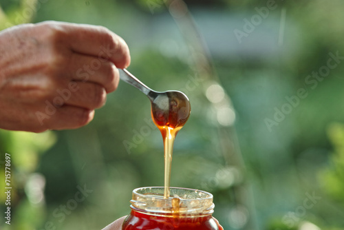 flowing honey dripping from the spoon into the preserving jar pot in the hand with green natural organic blurred bookeh background with effects