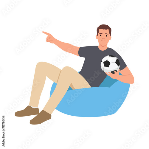 Young man support football sport team hold soccer ball watch tv live stream sit in bag chair point index finger aside.