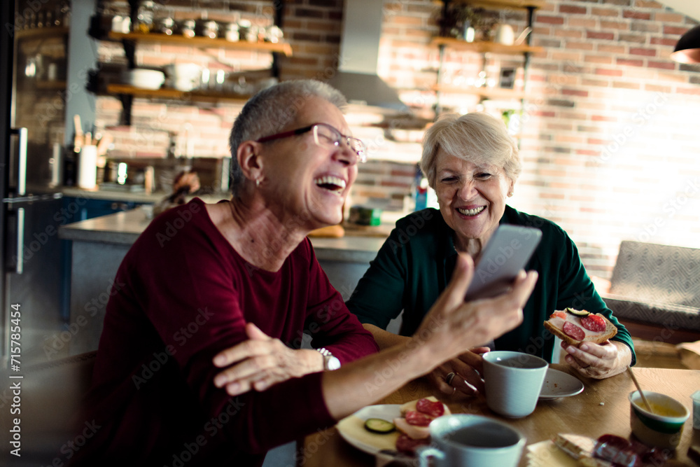 Two senior women having breakfast together and using smartphone at home Two senior women having breakfast together and using smartphone at home