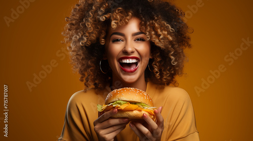  beautiful young woman eating hamburger isolated on yellow background. Woman hold big barbecue burger sandwich with hungry mouth happy screaming laughing on yellow background. Fast food concept.
