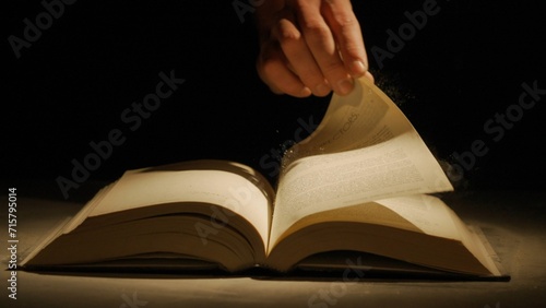 Studio shot of old retro hardcover book on dark background in warm light. Man holding page of old vintage book laying opened.