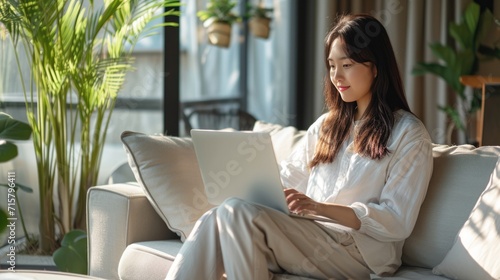 Asian young woman freelancer sitting on comfortable sofa browsing internet via laptop computer. Working from home concept. photo