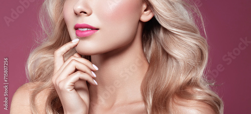 A woman with glossy blonde hair, pink lipstick, and manicured nails is posing, showcasing her flawless makeup and healthy skin...