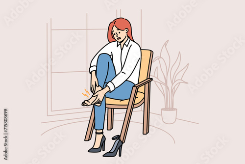 Woman with varicose veins feels pain in legs due to uncomfortable high-heeled shoes, sits on chair in apartment. Girl needs medicinal ointment to cure varicose veins or calluses on heel