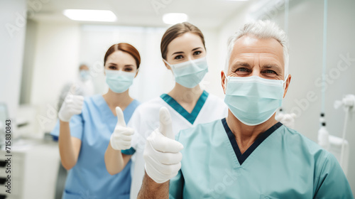 Healthcare professional in the foreground giving a thumbs-up and wearing a surgical mask, with a colleague in the background doing the same, both in a clinical setting.