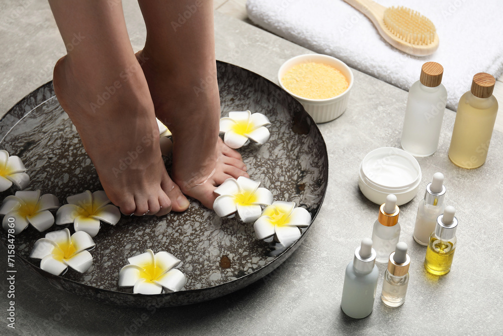 Woman soaking her feet in bowl with water and flowers on light grey floor, closeup. Spa treatment Woman soaking her feet in bowl with water and flowers on light grey floor, closeup. Spa treatment