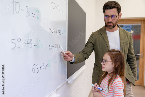 School teacher watching pupil writing on white board in classroom.
