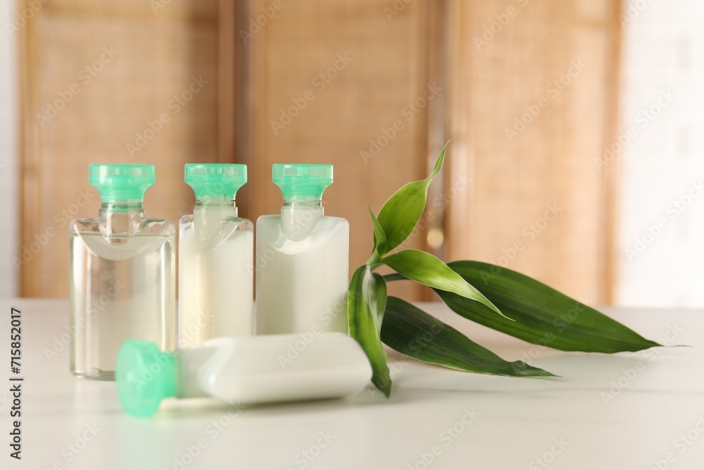 Mini bottles of cosmetic products and green branch on white table against blurred background. Space for text