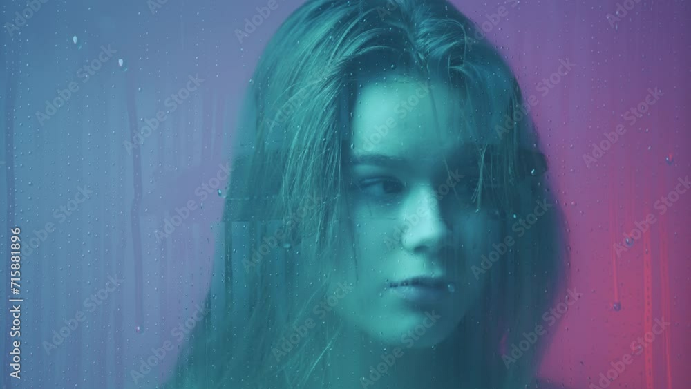 Portrait of female in neon light behind the glass window in steam and water drops. Girl with makeup looks around at camera through wet glass. Portrait of female in neon light behind the glass window in steam and water drops. Girl with makeup looks around at camera through wet glass.