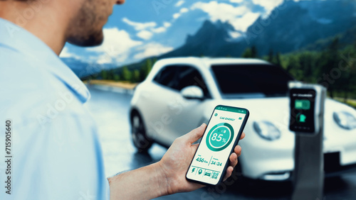 Businessman holding smartphone display battery status interface by smart EV mobile application while EV car recharging electricity from charging station with natural mountain outdoor background.Peruse