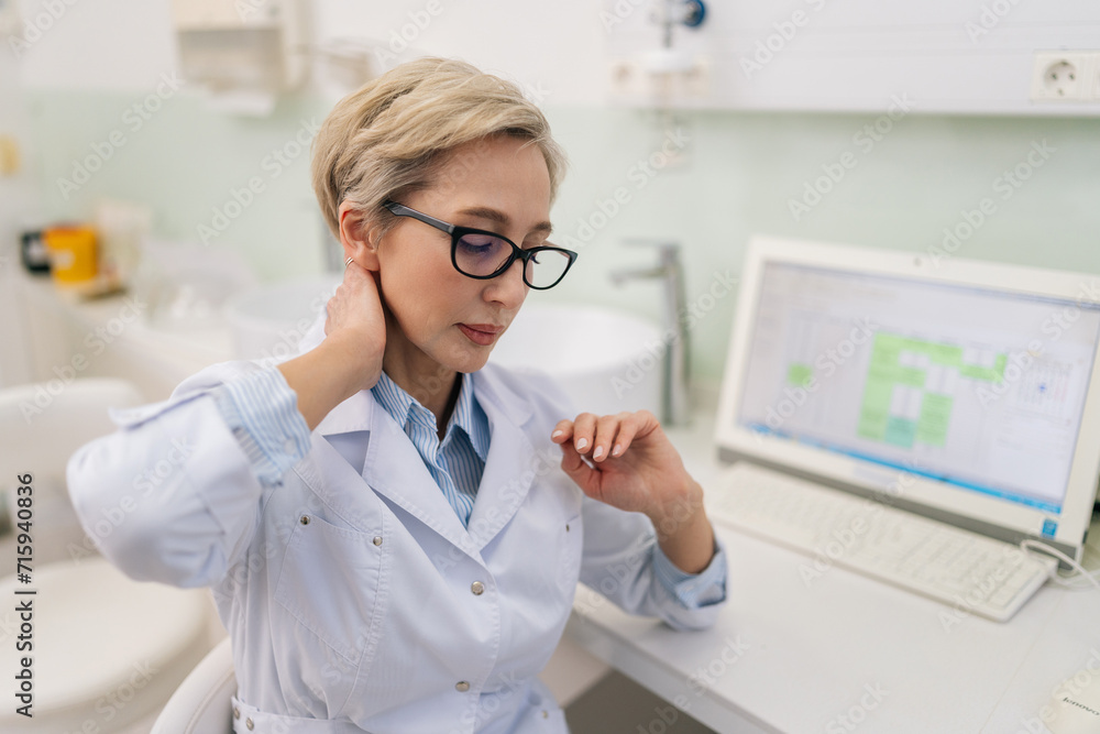 Portrait of tired middle-aged female doctor in eyeglasses having neck pain during working on laptop, suffering from injury at hospital. Mature woman medical expert holding painful area, spasm or ache. Portrait of tired middle-aged female doctor in eyeglasses having neck pain during working on laptop, suffering from injury at hospital. Mature woman medical expert holding painful area, spasm or ache.