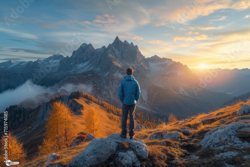 A solitary figure stands atop a rocky perch, gazing in awe at the majestic mountains looming before them, bathed in the warm hues of a breathtaking sunrise