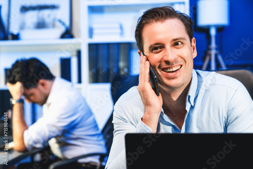 One of businessman calling on smartphone with smiling and happy face while coworker trying to make solutions with stressful feeling on blurred background. In casual comfy at workplace theme. Sellable.