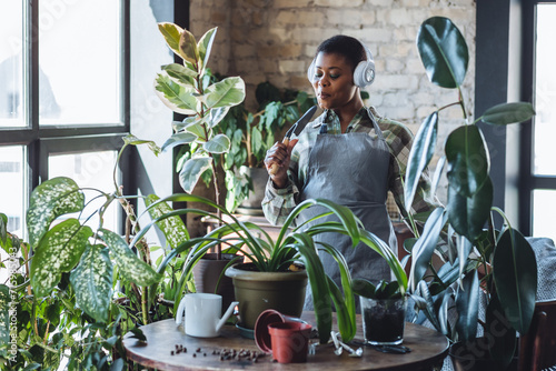 Happy young lady enjoying music, relaxing. Green trendy home interior. Carefree brunette African American woman listening to music, singing, using headphones, watering plants in living room.